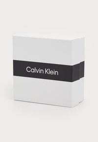 Scatola rettangolare bianca con una fascia nera su cui è presente la scritta "Calvin Klein" in testo bianco. Texture liscia, design minimalista, senza ulteriori dettagli.