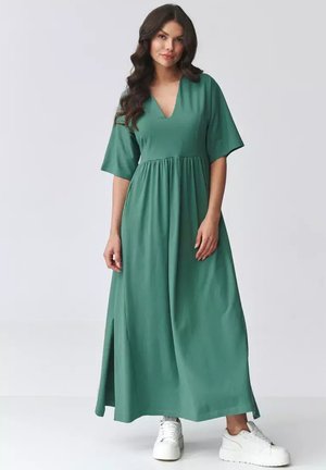 Kobieta stojąca, ubrana w długą sukienkę w kolorze zielonym z krótkimi rękawami i białe sneakersy na platformie, na tle jednolitego jasnego tła.