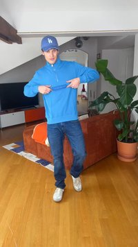 Νέος άνδρας με μπλε φούτερ και καπέλο δείχνει το λογότυπο ενώ στέκεται στο σαλόνι με πορτοκαλί καναπέ και μεγάλο γλαστρικό φυτό.