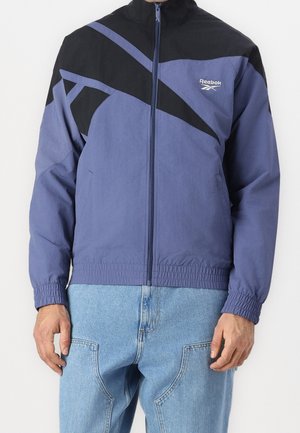 Fialová bunda Reebok s černými panelovými detaily, zapínáním na zip a elastickými manžetami. Hladká textura, lehký materiál.