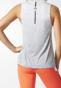 Harmaa hihaton urheilupaita, jossa on takaläppä ja musta pystysuora raita sekä logo. Yhdistetty oransseihin legginsseihin. Pehmeä, kevyt materiaali.