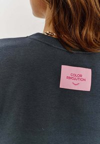 Tmavošedý mikina s rebrovaným výstrihom, s ružovou nálepkou, na ktorej je napísané "COLOR R3VOLUTION" červeným písmom.