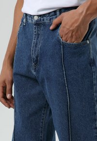 Jean en denim bleu foncé avec une coupe décontractée, cinq poches et des coutures contrastantes. Le tissu a une texture lisse avec un léger effet délavé.