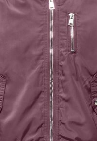 Veste bomber marron avec une texture lisse et brillante. Elle possède une fermeture éclair centrale argentée, deux poches latérales et une poche zippée supplémentaire sur la poitrine.