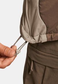 Beige och brun isolerad jacka med snörning och tryckknapp vid ärmen, med texturerad tyg och sydda detaljer.