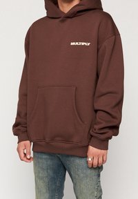 Felpa cappuccio marrone con tasca frontale, grande cappuccio e scritta "MULTIPLY" in bianco. Tessuto morbido, vestibilità ampia, polsini e orlo a coste. Design classico e casual.