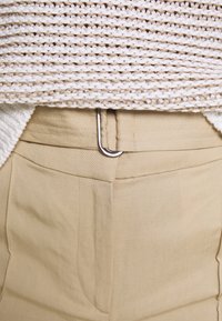 Beiga byxor har en slät textur, sidofickor och en silvermetallklämma på midjan, som kombineras med en strukturerad vit och beige stickad topp.
