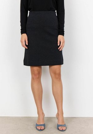 Femme portant un haut à manches longues noir et une jupe mi-longue à carreaux gris foncé, avec des talons ouverts bleus, debout contre un mur blanc.