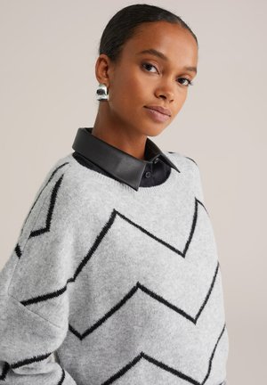 Young woman with pulled-back hair wearing silver earrings, a black collared shirt under a gray sweater with black zigzag stripes.