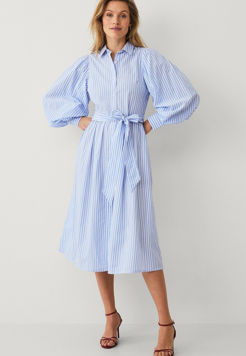 Kobieta ubrana w midi sukienkę w pionowe paski w jasnoniebieskim i białym kolorze, z bufkami na długich rękawach, przewiązaną paskiem w talii oraz bordowymi sandałami na obcasie.