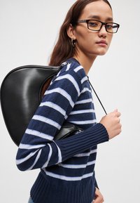 Μαύρο δερμάτινο σακίδιο με καμπυλωτό σχήμα, φορεμένο πάνω από ένα πουλόβερ με ριγωτή μπλε ναυτική και λευκή απόχρωση. Διαθέτει ριμπ μανσέτες και φερμουάρ κλεισίματος.