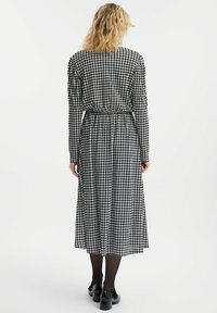 Abito lungo a scacchi bianco e nero con maniche lunghe, vita elasticizzata e gonna fluida, abbinato a scarpe nere e collant.