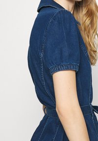 Kvinna som bär en mörkblå denimklänning med puffärmar och ett knytbälte i midjan, visar överkropp och arm.