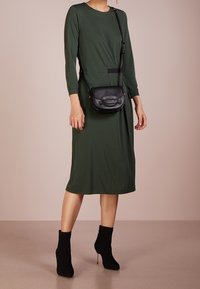 Robe verte olive à manches longues avec une coupe ample et un accent noir à la taille. Associée à des bottines noires et un petit sac bandoulière.