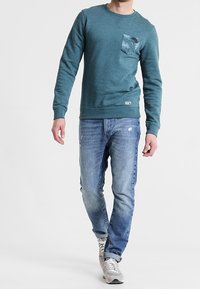 Felpa turchese con tasca sul petto a fantasia, abbinata a jeans azzurri chiari e sneakers grigio chiaro, presenta un design a vestibilità rilassata.