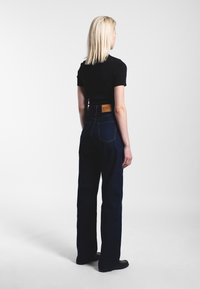 Vrouw met blond haar staat met haar rug naar de camera, draagt een zwarte top met korte mouwen, een hooggetailleerde donkerblauwe spijkerbroek en zwarte schoenen op een witte achtergrond.