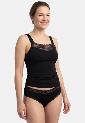 Donna sorridente che indossa un top nero con bordi in pizzo e biancheria intima abbinata con bordi in pizzo, in piedi davanti a uno sfondo semplice.