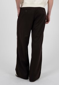 Vue de dos d'une personne portant un pantalon large marron foncé avec des poches arrière boutonnées et des chaussures de couleur claire sur un fond neutre.