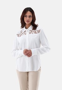Camicia bianca con bottoni, maniche lunghe e colletto, caratterizzata da un motivo in pizzo sul petto. Tessuto morbido, vestibilità rilassata e polsini abbottonati.