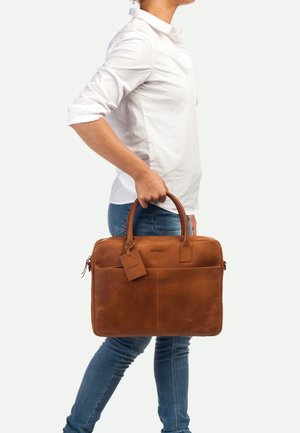 Malette en cuir brun avec deux poignées supérieures, surface texturée, poche zippée à l'avant et étiquette amovible, portée par une personne en tenue décontractée.