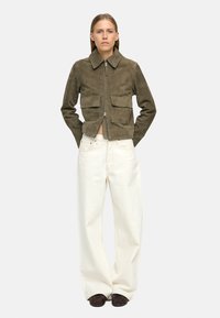 Zielona oliwkowa kurtka z zamszu z zamkiem z przodu, dwiema kieszeniami z przodu i kołnierzem; zestawiona z białymi dżinsami o szerokich nogawkach i ciemnym obuwiem.