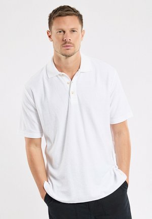 Homme aux cheveux courts portant un polo blanc à manches courtes et un pantalon noir, debout les mains dans les poches devant un fond blanc.