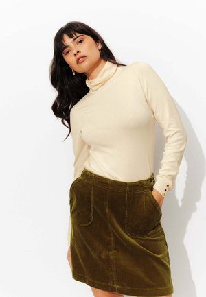 Top beige à col roulé à manches longues, associé à une jupe en velours côtelé vert olive dotée de poches et de boutons décoratifs.