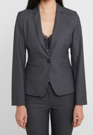 Mujer que lleva una americana entallada de color gris oscuro con un botón, combinada con pantalones a juego y una blusa de encaje negra debajo.