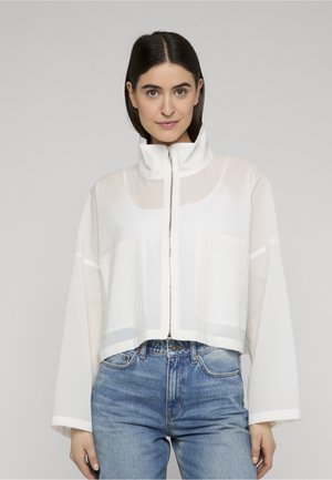 Kvinde iført en semi-gennemsigtig hvid lynlåstrøje med høj krave og blå jeans, der kigger lige frem mod en neutral baggrund.