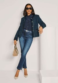 Frau in dunkler Sonnenbrille, die eine navyblaue strukturierte Jacke, Bluse, knöchellange Jeans, beige Absatzschuhe trägt und eine beige Handtasche hält.