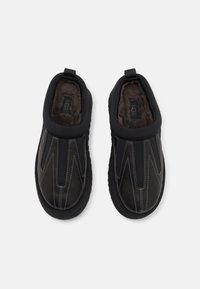 Paire de chaussures noires sans lacets avec des coutures blanches visibles et une doublure en peluche sombre, vue de dessus sur un fond clair.