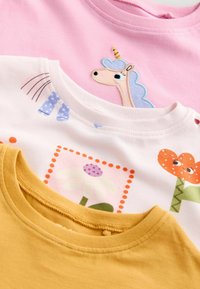 Trois chemises à manches courtes : une rose avec une licorne, une blanche avec un motif floral, et une jaune moutarde. En coton doux, conception col rond et motifs ludiques.
