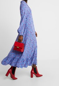 Modré květinové šaty s dlouhými rukávy a volánkovým lemem, doplněné červenými kotníkovými botami a odpovídající kabelkou se zlatým zapínáním a hroty.