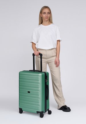 Donna in camicia bianca e pantaloni beige in piedi con una valigia rigida verde con manico estensibile e quattro ruote su sfondo bianco.