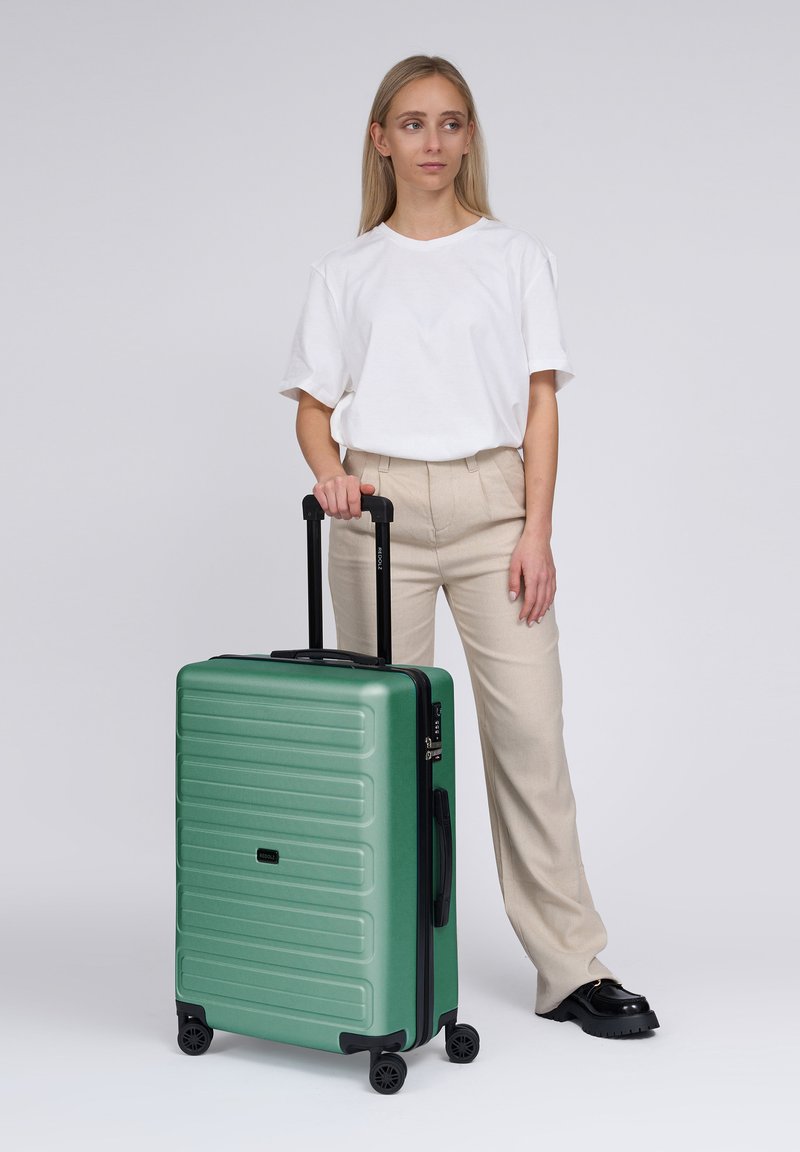 Mujer con camisa blanca y pantalones beiges de pie junto a una maleta rígida verde con asa extensible y cuatro ruedas sobre fondo blanco.