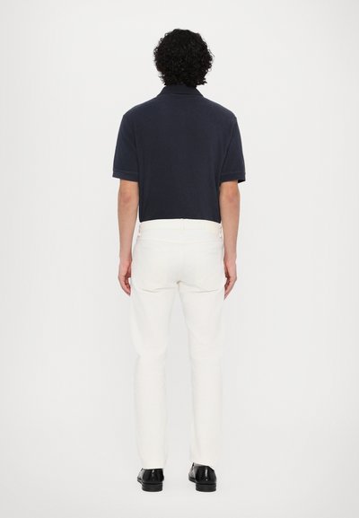 Marinblå kortärmad polo med enkel krage, ihop med slim-fit vita byxor och svarta skor, sedd bakifrån.
