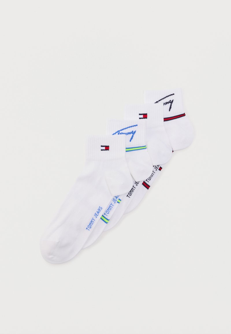 Τέσσερα ζευγάρια λευκές κάλτσες αστραγάλου με το λογότυπο Tommy Jeans και πολύχρωμα λογότυπα στις μανσέτες και στα πλαινά, τοποθετημένα διαγώνια σε λευκό φόντο.