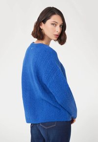 Pullover blu lavorato a maglia con un motivo a treccia testurizzato, scollatura rotonda, spalle cadute e orlo a costine. Indossato sopra jeans in denim scuro.
