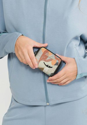 Person in hellblauer Jacke und Hose, die eine kleine, bunte Geldbörse mit abstraktem Katzenkopf-Design und Reißverschluss hält.