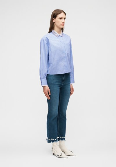 Γυναίκα που στέκεται φορώντας ένα ανοιχτό μπλε ριγέ μπλουζάκι με λουλουδάτο γιακά, τζιν παντελόνια cropped με λουλουδάτη διακόσμηση, λευκές κάλτσες και λευκά μυτερά παπούτσια.