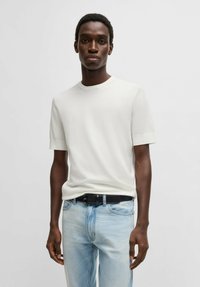 Hvid kortærmet t-shirt lavet af blødt stof, med rund hals design, båret med lyseblå jeans og et sort bælte. Glat tekstur med minimal detalje.