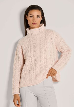 Frau in einem hellrosa Grobstrickrollkragenpullover und hellgrauen Hosen, die vor einem einfarbigen beigen Hintergrund posiert.