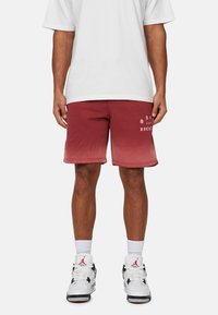 Pantaloni corti con sfumature di rosso e dettagli in logo bianco, tessuto leggero, cintura elastica e vestibilità comoda, abbinati a sneakers bianco e nere.