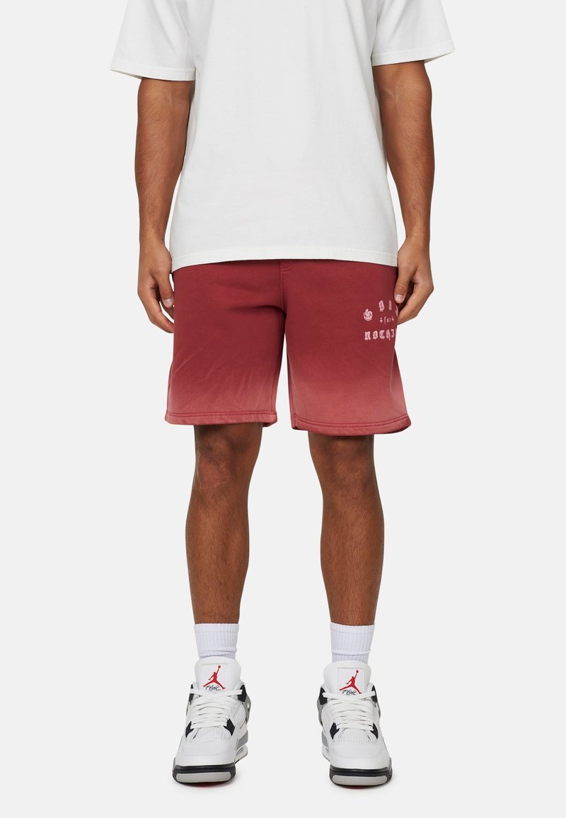 Pantaloni corti con sfumature di rosso e dettagli in logo bianco, tessuto leggero, cintura elastica e vestibilità comoda, abbinati a sneakers bianco e nere.