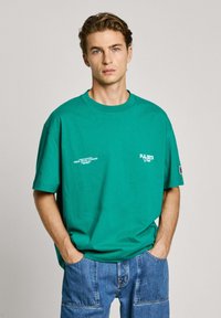 Zöld, oversized pamut póló, kerek nyakkal, rövid ujjakkal, és nyomtatott logó részletekkel a mellkason és az oldalon. Farmernadrággal párosítva.