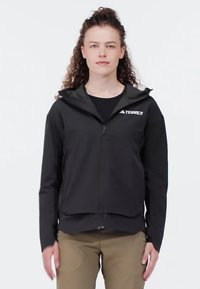 Svart zip-up jacka med huva, tillverkad av ett lätt material. Har en logotyp på övre höger sida och en strömlinjeformad design.