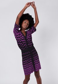 Abito a zigzag lilla e nero con scollo a V, maniche corte e dettagli a bottoni lungo la parte anteriore. La vita a coste accentua la silhouette.