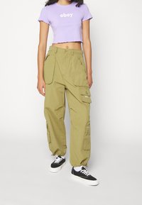 Světle fialový zkrácený tričko s nápisem "obey"; olivově zelené cargo kalhoty s bočními kapsami; černé tenisky s bílými podrážkami a tkaničkami.