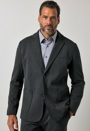 Uomo con barba corta che indossa una giacca da completo grigio scuro e una camicia grigio chiaro abbottonata su uno sfondo chiaro uniforme.
