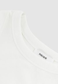 Valkoinen puuvillapaita, jossa on pyöreä pääntie ja ommeltuja yksityiskohtia; tarrassa lukee "REISS." Liukas pinta, yksinkertainen muotoilu.